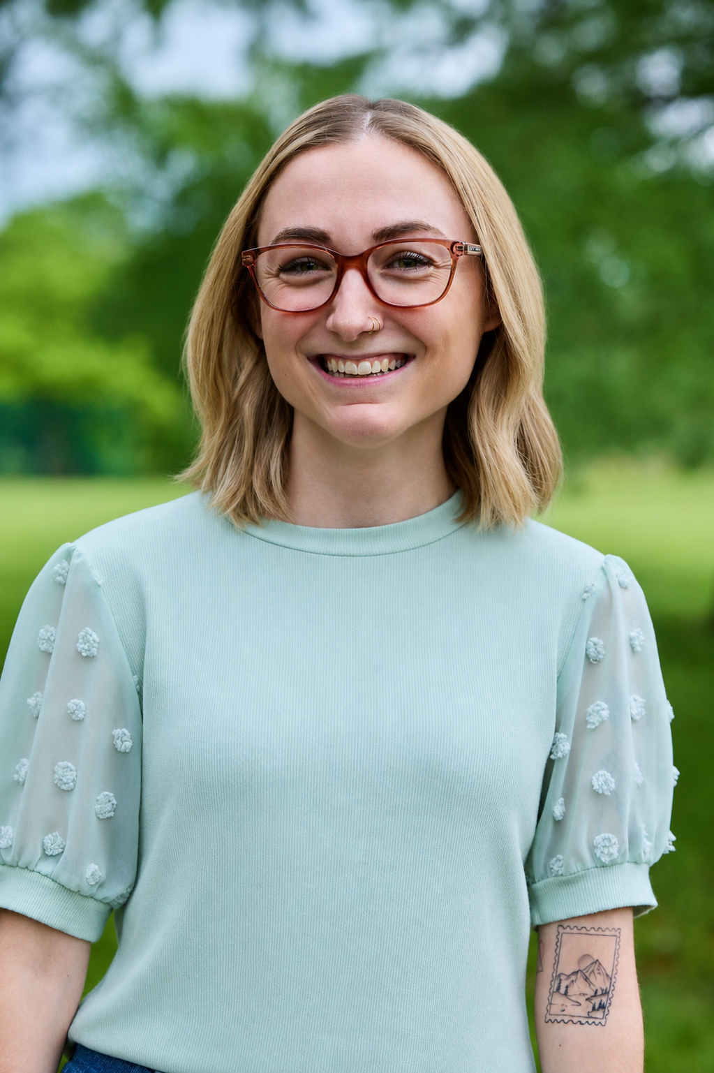 Danielle Graff Low-Res - Change, Inc. St. Louis Counseling Smiling woman with shoulder-length blond hair and red glasses outdoors, wearing a mint-green top with textured puff sleeves; a small tattoo is visible on her forearm.
