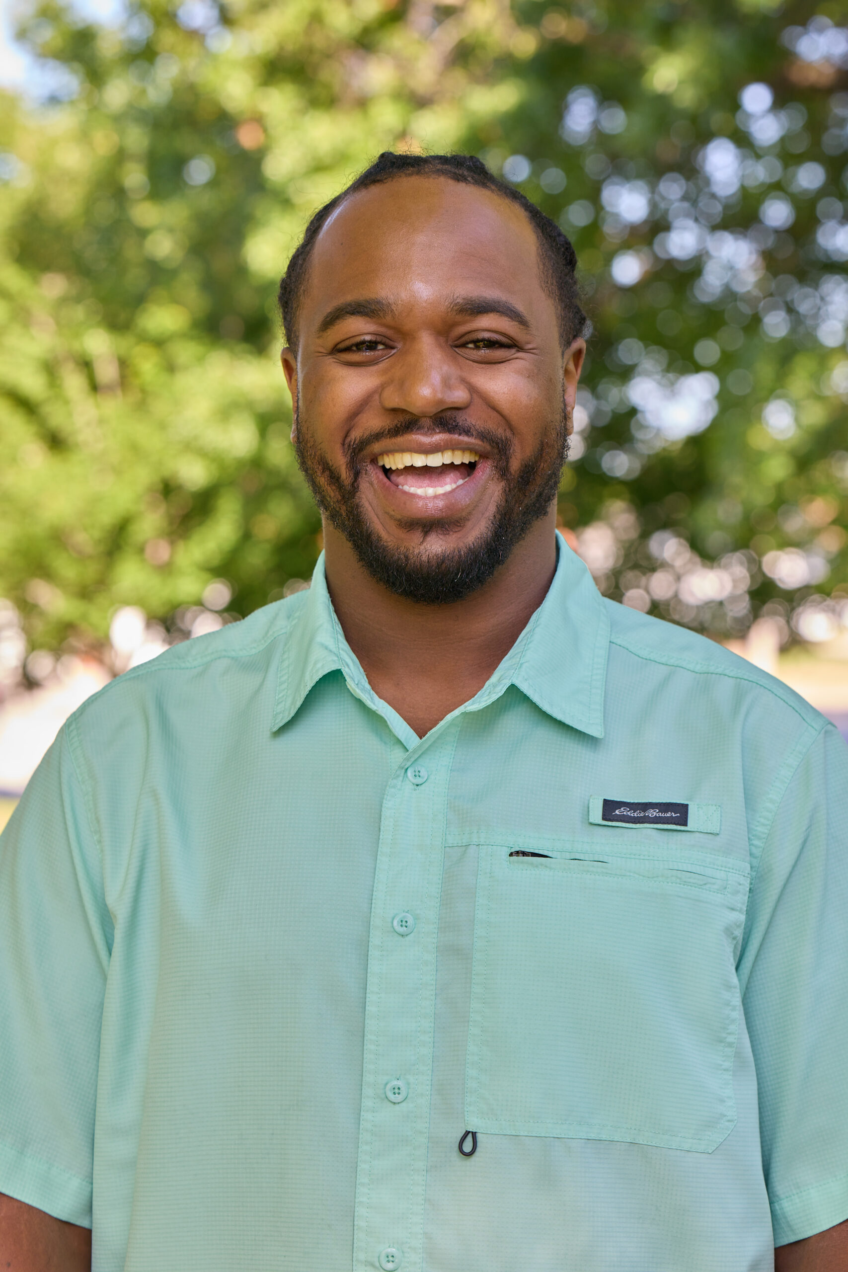 Tre Davis Low Res Smiling man with a beard outdoors, wearing a mint green button-down shirt, with a blurred green foliage background