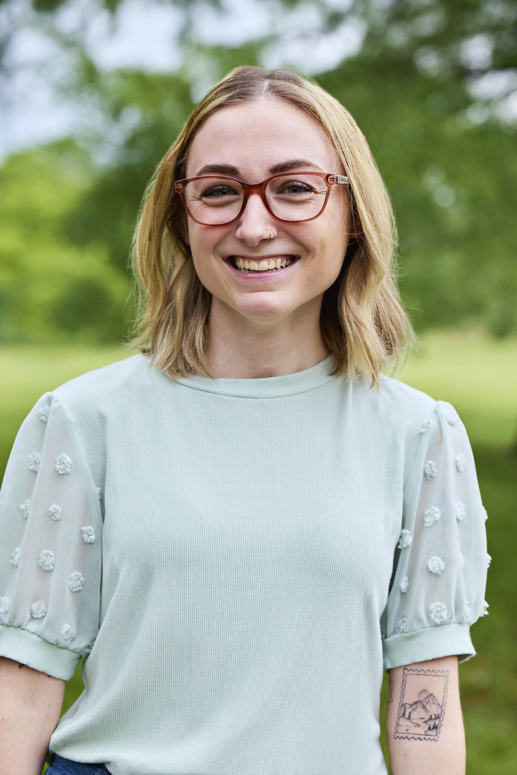 Danielle Graff Hi-Res - Change, Inc. St. Louis Counseling Smiling woman outdoors wearing red-brown glasses and a light mint-green top with sheer, embroidered sleeves; a tattoo is visible on her forearm.